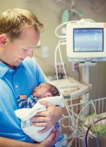 Young smiling father holding her Premature newborn baby who is being treated in the hospital. With love and tenderness he holds his baby close in a hospital room next to a vital signs monitor