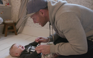 Kneeling dad wearing a blue cap and grey hoodie holding hands with his baby, who is lying on the floor. They are making clear eye contact; the dad is smiling at his baby and the baby looks engaged and open-mouthed in response.